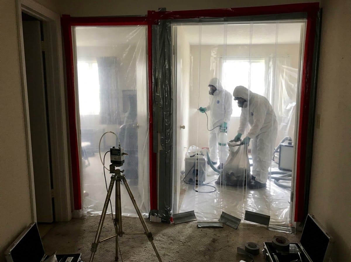 A room sealed with plastic sheeting for asbestos removal, with an air monitoring pump outside the entrance